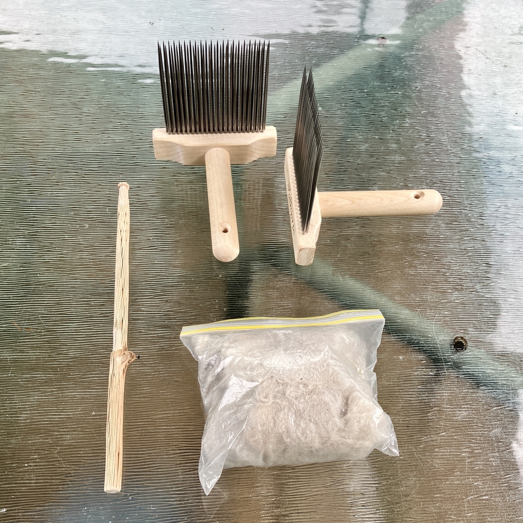 Two wool combs, a hand distaff, and a small plastic bag full of beige fibre sit on a glass table.
