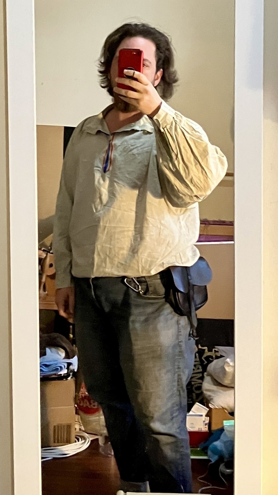 A selfie of a white person taken in a long, narrow mirror. Their face is obscured by their phone. They are wearing a light green pirate shirt with rainbow trim on the neck slit. It is tucked into mid-blue denim jeans  and blouses at the belt line. There is a black leather kidney pouch at their belt. End ID. 