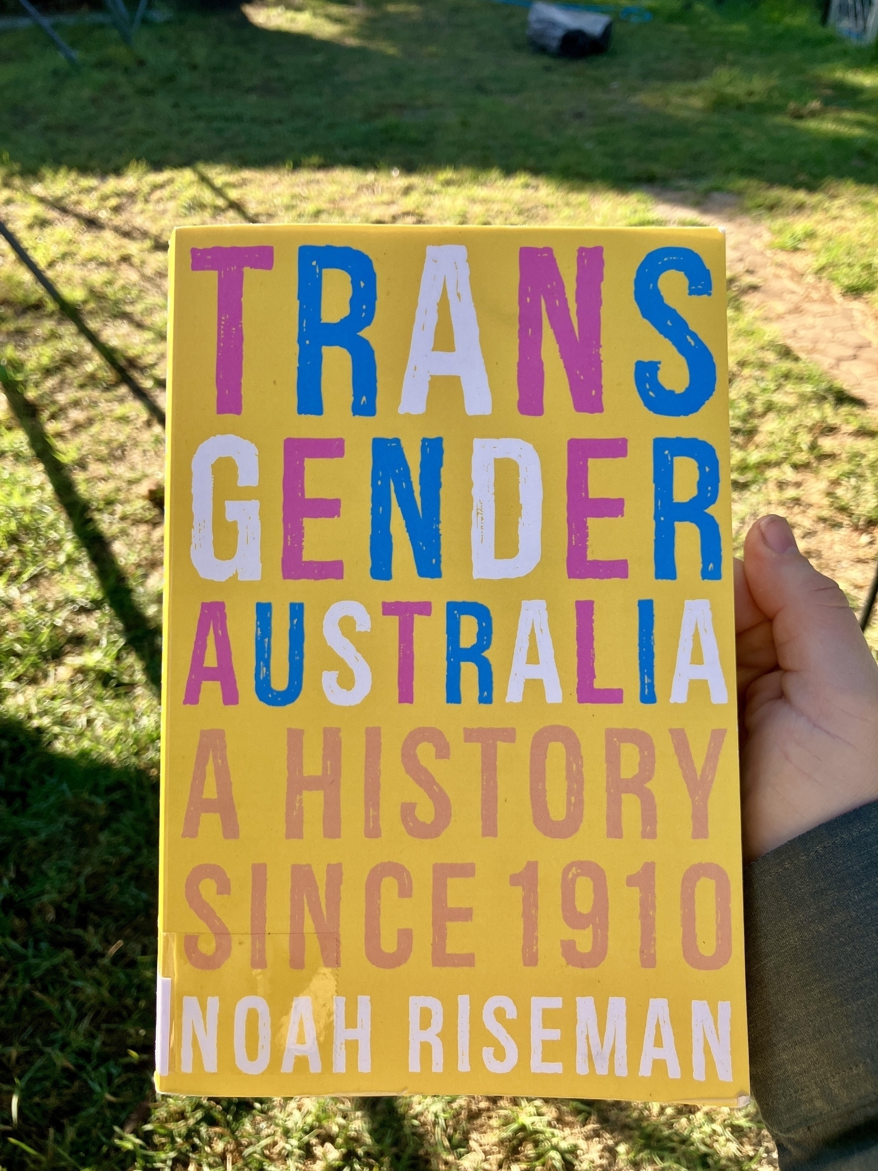 The cover of a book titled “Transgender Australia: A History Since 1910” by Noah Riseman. The words “Transgender Australia” are in the colours of the trans pride flag. The subtitle is in dark pink and the author’s name is white. The cover background is bright yellow. 