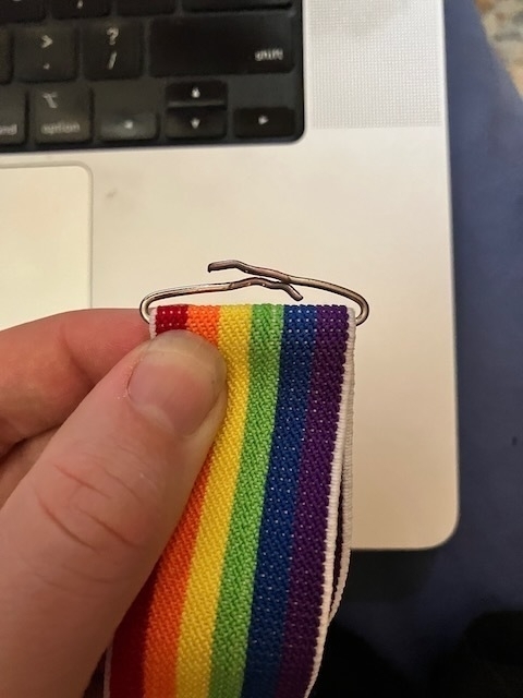 The end of a pair of clip-on, rainbow elastic suspenders. The wire that attached it to the elastic has been bent so that the ends that were holding the clip in place overlap each other. End ID.