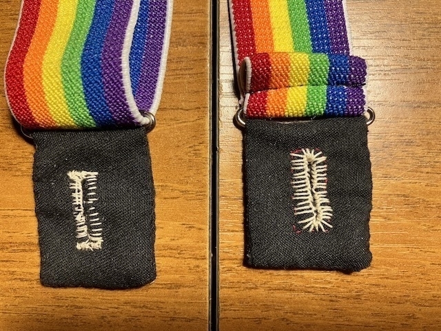 The ends of two suspender straps sit side by side on a wooden table. They have had tabs of black cloth sewn onto the ends. There is a hand-sewn buttonhole done in thick cream thread on each cloth tab. End ID.