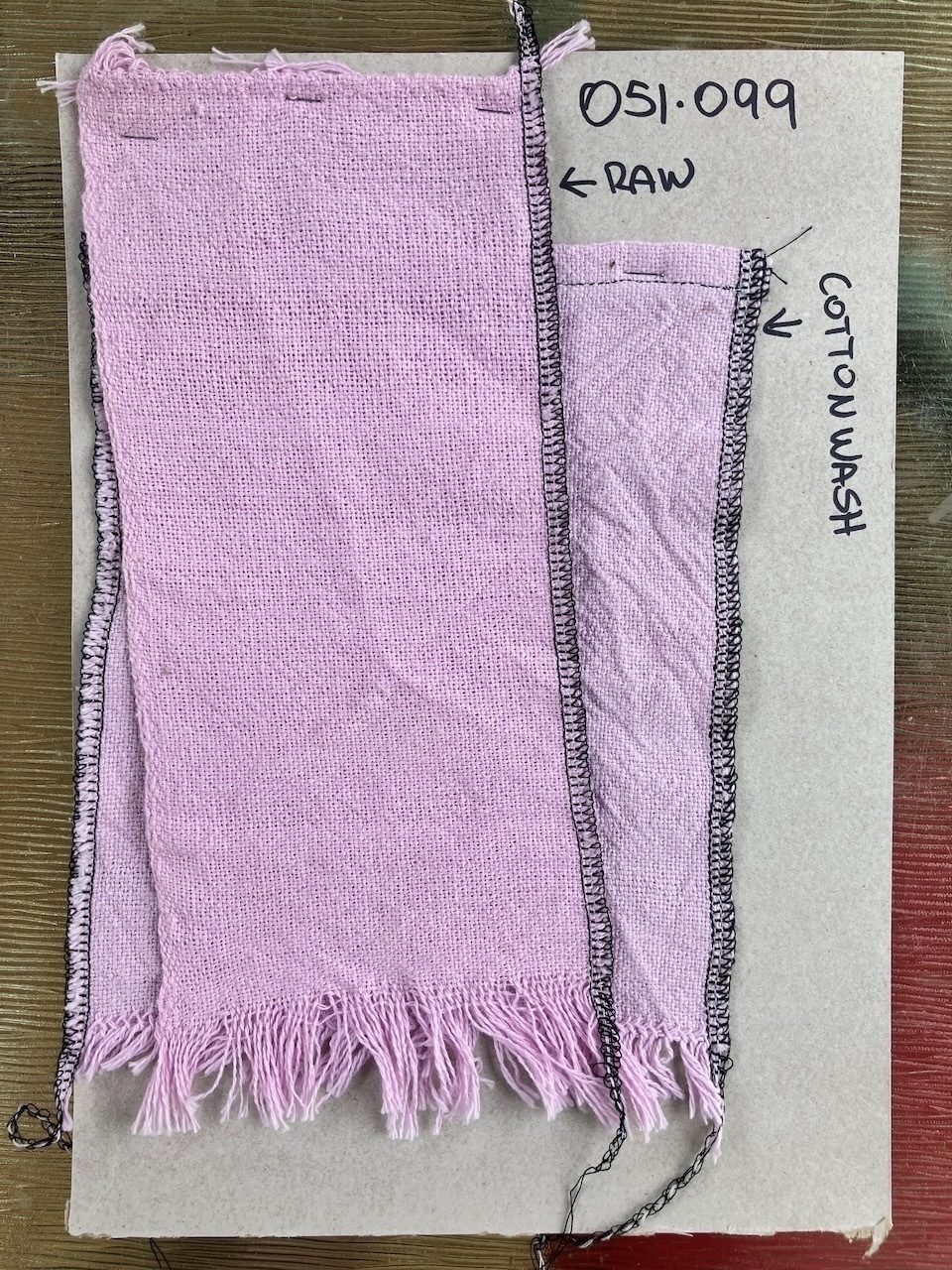 A piece of cardboard about the size of an A4 sheet of paper. There is a project number written in black Sharpie in the top right corner. The cardboard has two pieces of light pink, handwoven cloth stapled to it, one above the other. The one at the top is labelled 'raw'. The one underneath is labelled 'cotton wash'. End ID.